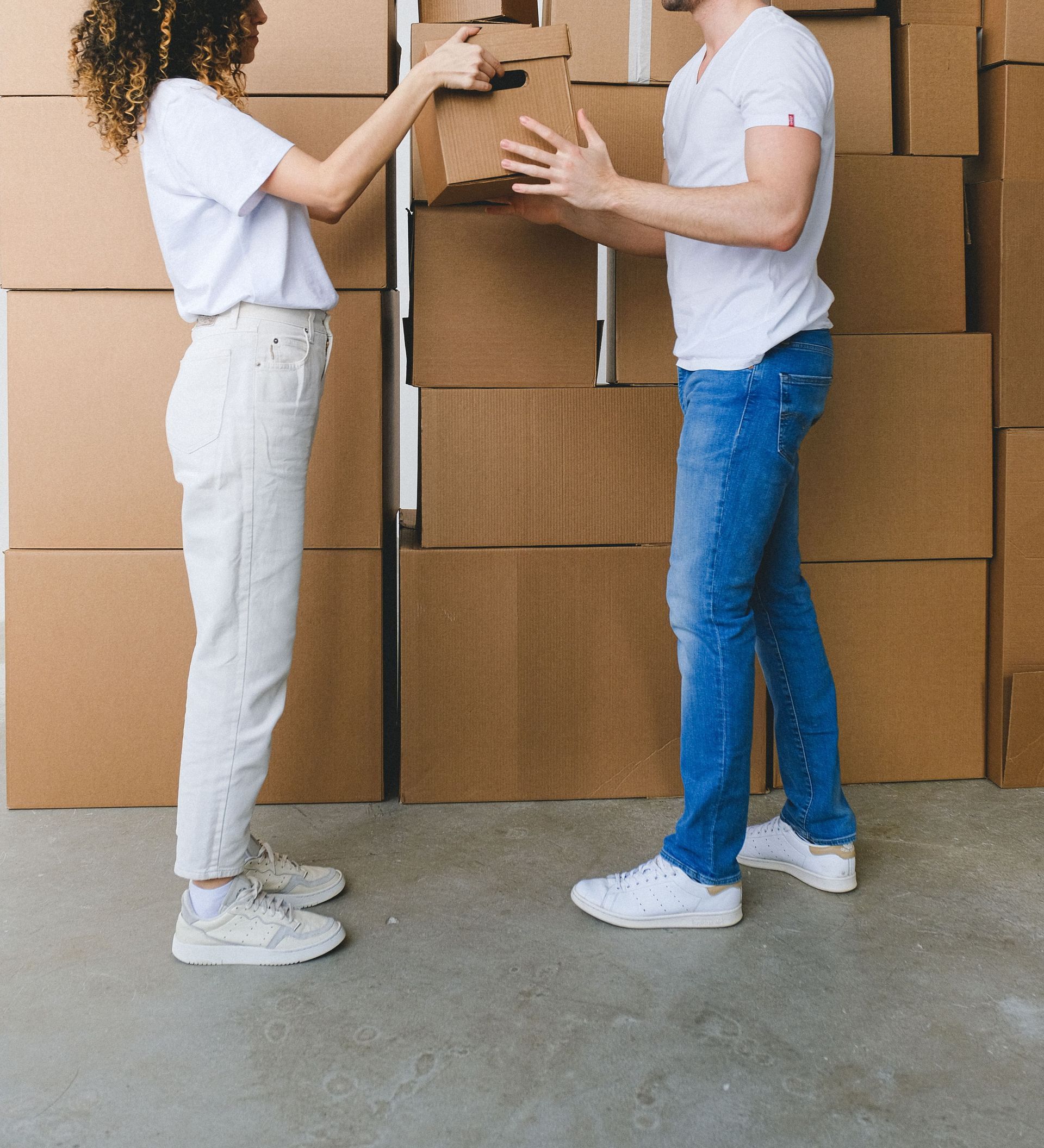 Side view of young lady with curly hair in casual clothes passing cardboard box to boyfriend during moving in new apartment in daylight