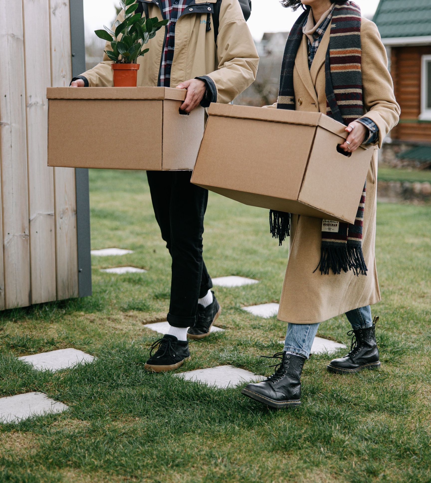 Happy couple carrying boxes and a plant while moving into their new home on a grassy lawn.